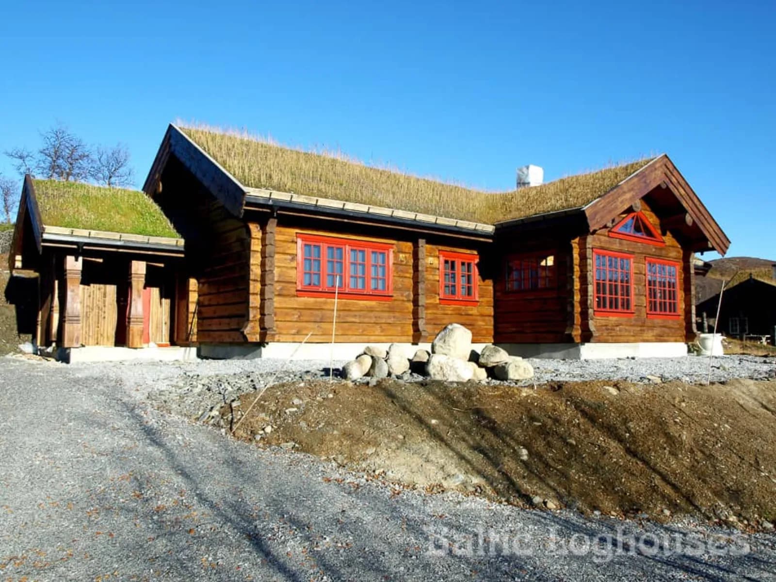 Ühekorruseline murukatusega palkmaja eredas päikesevalguses, punased aknaraamid, kivihunnik ja kruusatee ees, pinnas veel osaliselt paljas.