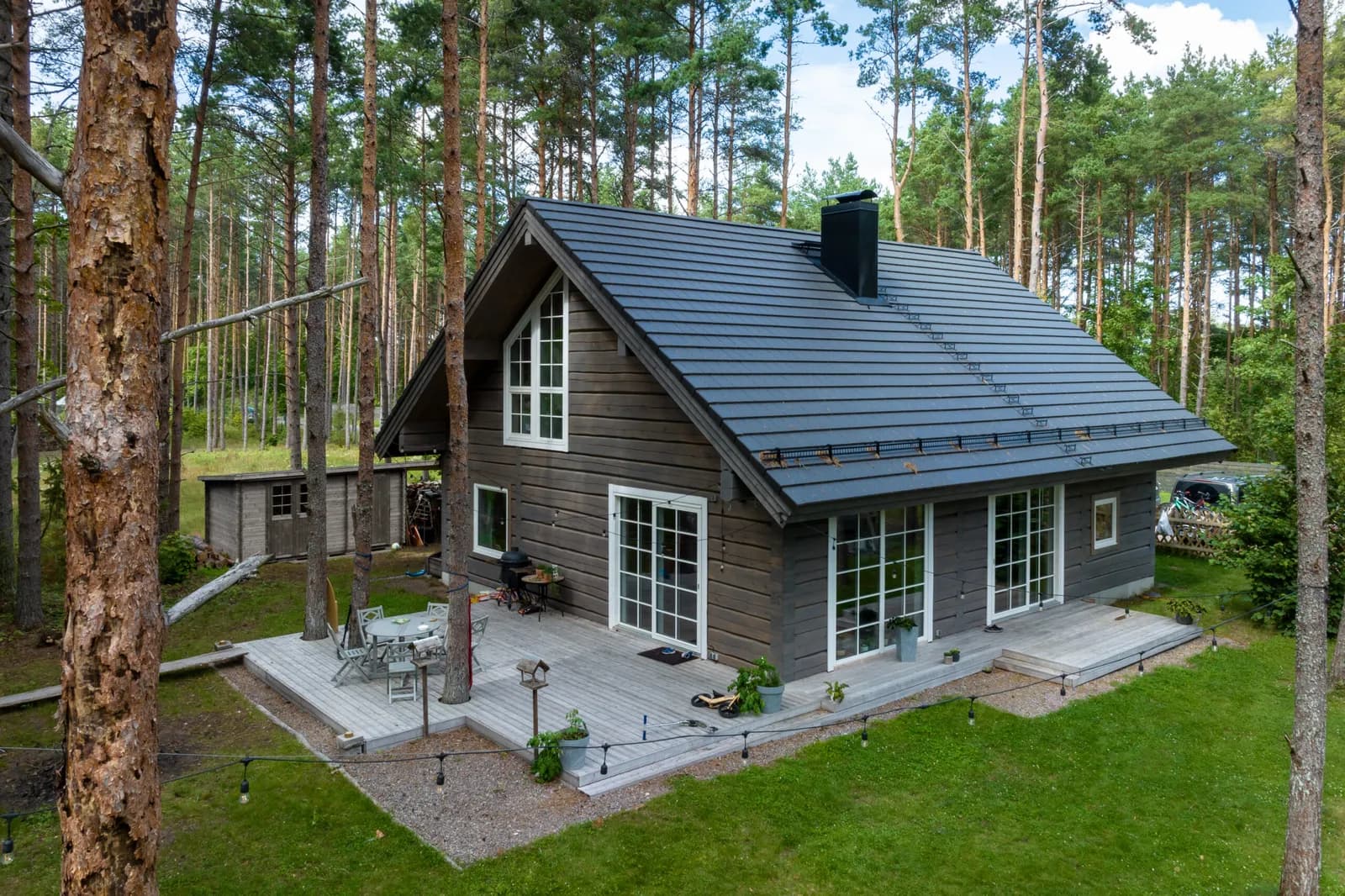 Palkmaja välisvaade, tumeda katusega ja tumepruuni puitvoodris fassaadiga männimetsas, terrass välimööbliga. Töö tegi Hobbiton.