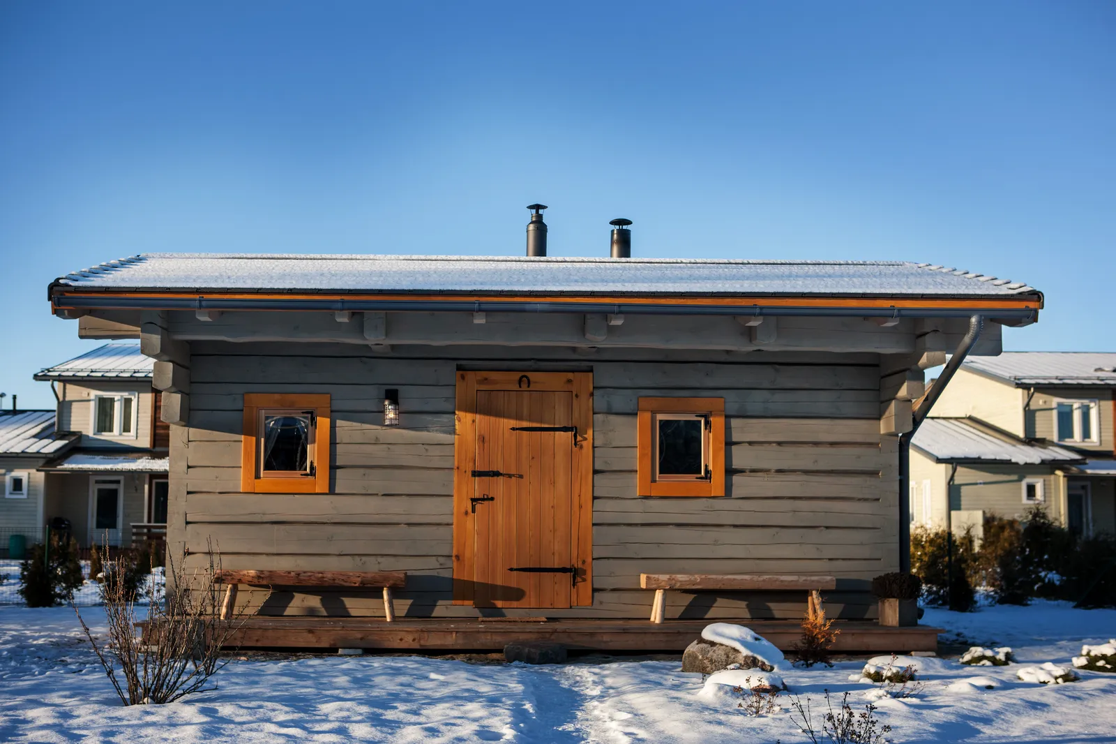 Väikese hallika palkhoone välisvaade lumisel krundil, oranžide aknaraamidega ja kahe musta korstnatoruga katusel, naabermajad taustal. Töö tegi Hobbiton.