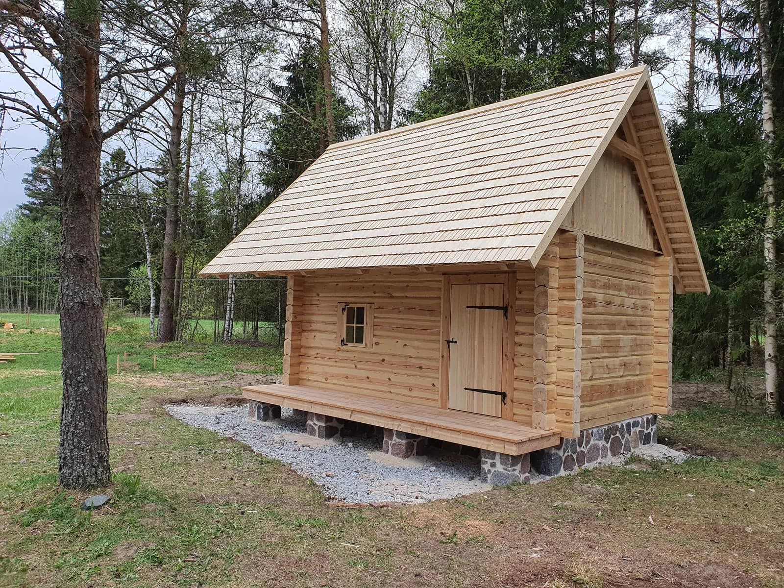 Väike loodusliku viimistlusega palksaun kivivundamendil, laastukatus, metsaserv taustal. Töö tegi Hobbiton.