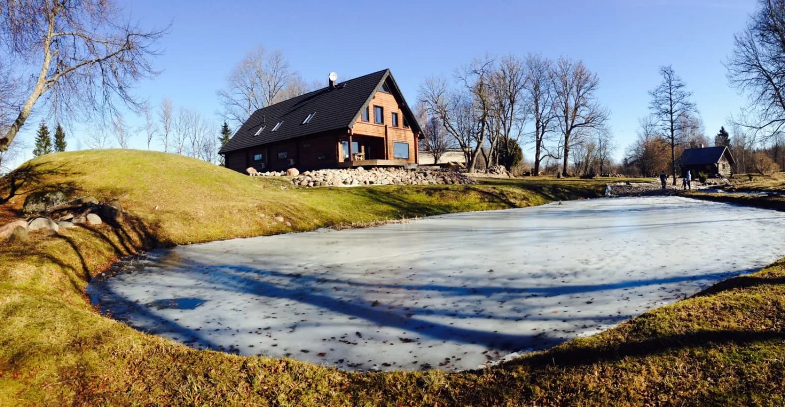 Tumedaks lasuritud palkmaja tiigi ääres, viilkatusel (mitte kelpkatusel) kaks katuseakent, jäätunud tiik, pruunikas muru ja lehtedeta puud, lund maas ei ole. Töö tegi L.L. House.