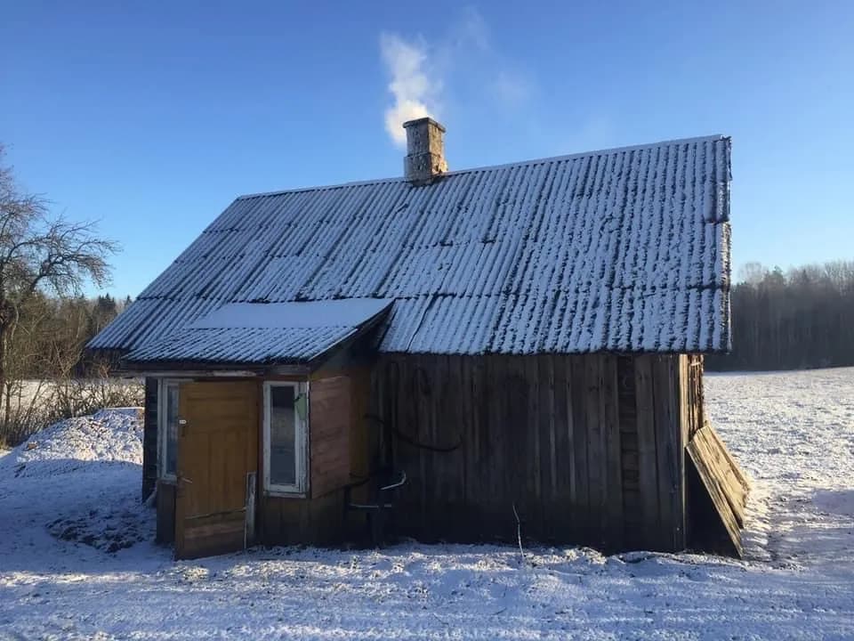 Väike kulunud puitelamu talvel lumisel põllul: madala kaldega laineplekk-katus, tumedad voodrilauad, korstnast tõuseb suitsu, vasakul lisatud varikatusega sissepääs.
