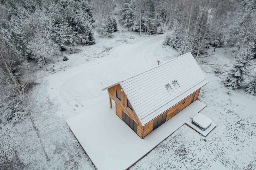 Droonifoto üksikust puitkatusega hoonest lumises metsalagendikus, teeviit ja sõiduki jäljed lumel. Töö tegi Eesti Käsitöö Palkmajad.